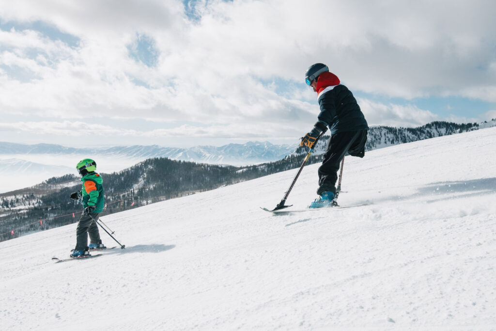 Adaptive skier with ski pole attachments skiing down a run with a young kid