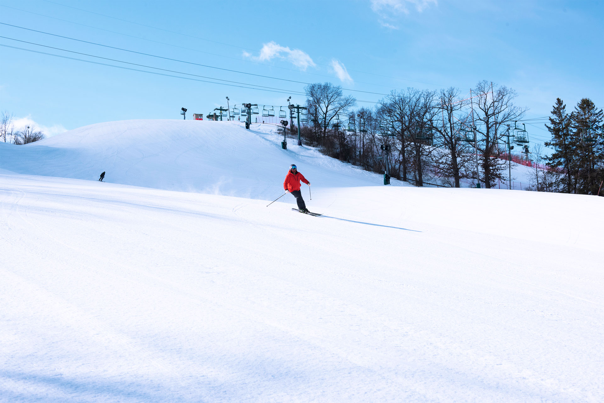 Skier in a red jacked skiing down a groomed run with a lift in a background