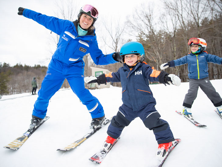Ski instructor and 2 your children smiling and posing for a photo with their arms out to the side like they're flying