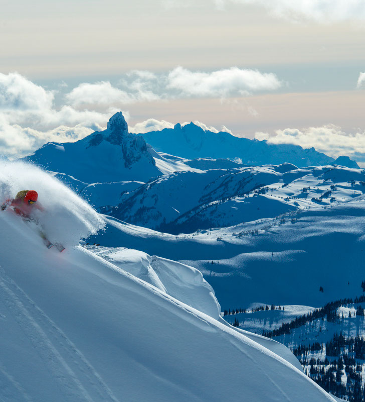 Skier in red jacket carving a turn in powder with scenic mountain views of Whistler Blackcomb in the background