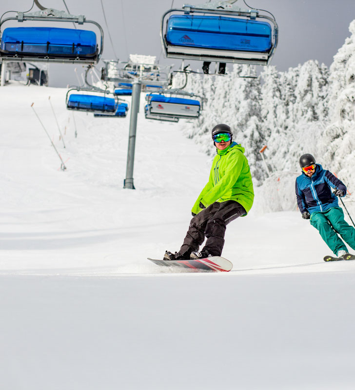 Skier and Rider Make Turns Under Chairlift on Snowy Day at Mount