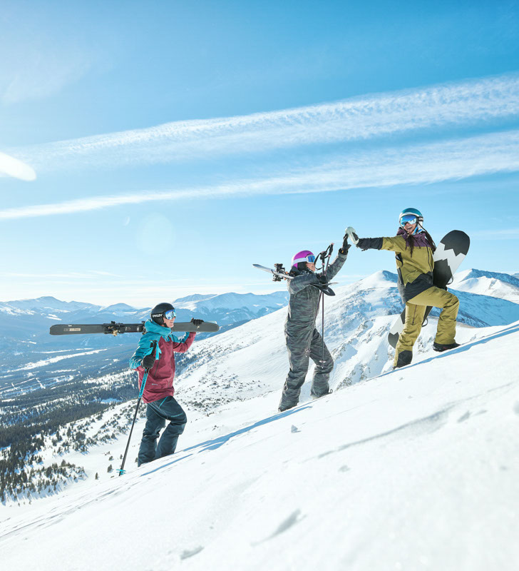 image of 2 skiers and a snowboarder hiking up to a run at breck on a sunny day. One skier high fives the snowboarder