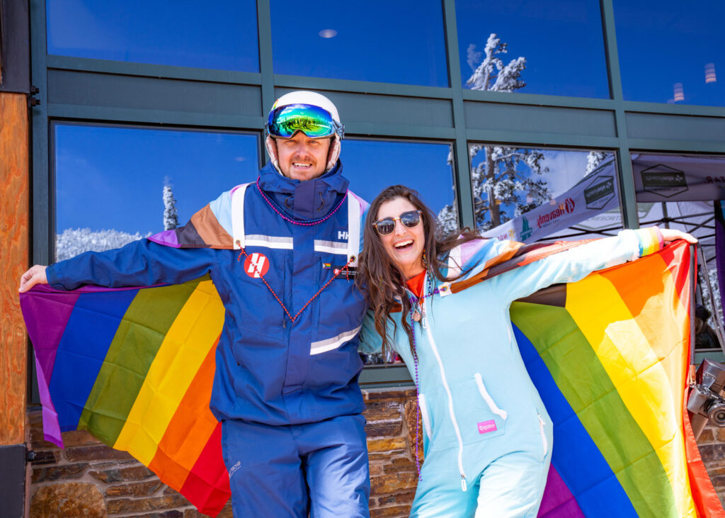 Heavenly employees smiling wearing pride flags with their arms outstretched in front of a resort building with glass windows