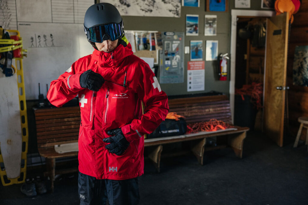 Man wearing a ski helmet and zipping a ski patrol jacket inside of a ski patrol work shop