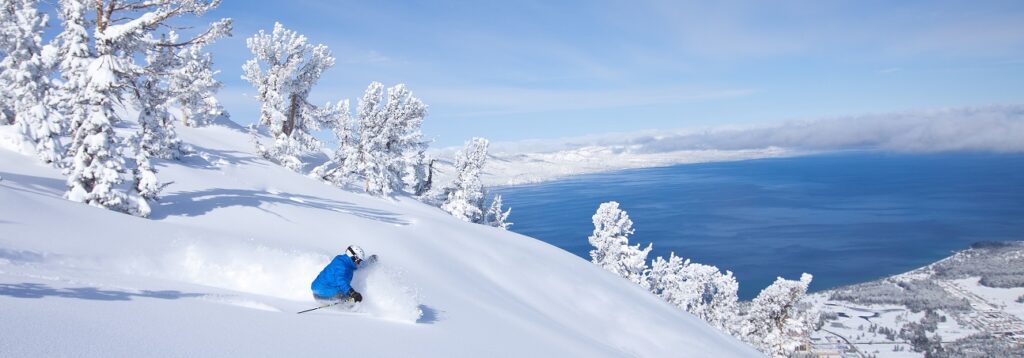Skier in blue jacket skiing down a powder run at heavenly with lake tahoe in the background