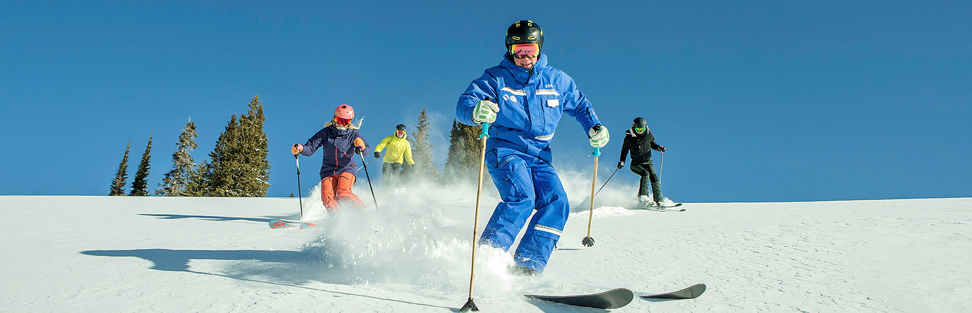 A ski instructor and three skiers skiing down a snowy ski slope with trees and a clear blue sky in the background